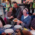 A group of people of various ages, all wearing wireless headphones, gather around a table set with white plates and glasses. A young girl serves soup from a bowl to an older man seated across from her. The mood is communal and lively.