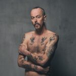 A shirtless man with tattoos covering his arms, chest, and stomach stands against a plain, gray background. He has a mustache and goatee, and his hair is shaved on the sides with long hair on top, tied back. He crosses his arms over his torso.