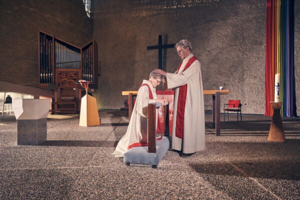 Een persoon in een religieus gewaad knielt bij een altaar, terwijl een andere persoon in een soortgelijk gewaad staat en zijn handen op zijn hoofd legt om te zegenen. Dit alles gebeurt in een kerk met een kruis en kleurrijke linten op de achtergrond.