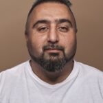 Een man van middelbare leeftijd met kort donker haar, een getrimde baard en snor draagt een wit T-shirt en kijkt recht in de camera tegen een effen bruine achtergrond.