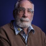 Een oudere man met een witte baard en bril draagt een bruine trui met rits over een blauw geruit overhemd. Hij heeft een geel lint op zijn kraag gespeld en een kippah op zijn hoofd, poserend tegen een donkerblauwe achtergrond.