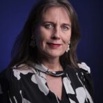 Een vrouw met lang bruin haar die een zwart-witte bloemenblouse en bloemvormige oorbellen draagt, staat tegen een donkerblauwe achtergrond en kijkt met een lichte glimlach naar de camera.