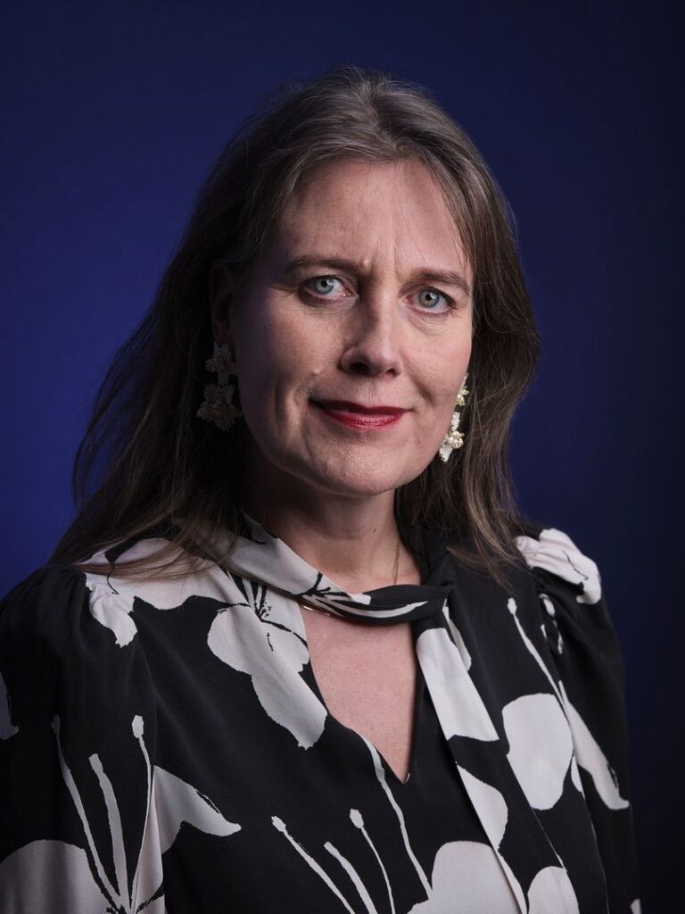 Een vrouw met lang bruin haar die een zwart-witte bloemenblouse en bloemvormige oorbellen draagt, staat tegen een donkerblauwe achtergrond en kijkt met een lichte glimlach naar de camera.