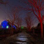 Een verlaten huis verlicht met blauwe lampjes staat aan het einde van een met bomen omzoomd pad. De bomen gloeien rood op en een gloeiende lichtbol zweeft nabij het huis onder een nachtelijke hemel.