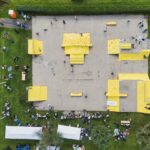 Luchtfoto van een betonnen skatepark met felgele schansen en obstakels, omgeven door groen gras. Mensen skaten, wandelen en verzamelen zich rond witte tenten en banken aan de rand van het park.