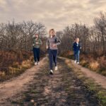 Drie mensen joggen op een onverhard pad door een bos met kale bomen en een bewolkte hemel, ze dragen sportkleding en hardloopschoenen.