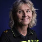 Een glimlachende oudere vrouw met blond haar draagt een donker politie-uniform met gele strepen en insignes en staat voor een effen donkerblauwe achtergrond.