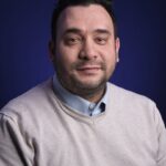 Een man met kort donker haar en een getrimde baard draagt een lichtbeige trui over een blauw overhemd. Hij poseert tegen een donkerblauwe achtergrond en kijkt naar de camera met een neutrale uitdrukking.