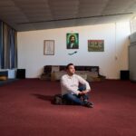Een man zit in kleermakerszit op een rood tapijt in een ruime, zonovergoten kamer met lage stoelen langs de muren. Achter hem hangen ingelijste portretten en een groen religieus symbool op een witte muur.