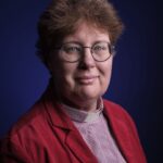 Een persoon met kort, krullend bruin haar, een bril en een lichte huid draagt een rood jasje, een geruit overhemd en een administratieve kraag. Ze poseren tegen een donkerblauwe achtergrond, kijken naar de camera en glimlachen zachtjes.