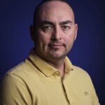 Een man met kort donker haar en een lichte huid en een geel overhemd met kraag poseert tegen een donkerblauwe achtergrond en kijkt met een neutrale uitdrukking recht in de camera.