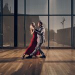 Een vrouw in een rode jurk en een man in een grijs pak dansen dicht op elkaar op een gepolijste houten vloer in een zonovergoten kamer met hoge ramen en een potplant in de hoek.