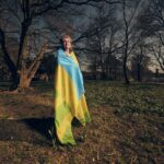 Een oudere persoon staat buiten in een park, gehuld in een grote blauwe en gele vlag, met kale bomen en een bewolkte lucht op de achtergrond.