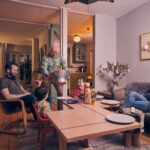 Een gezin in een gezellige woonkamer: twee volwassenen zitten op stoelen, een vrouw zit op een bank, twee kinderen zitten aan tafel en een oudere man schenkt popcorn in een kom. De kamer heeft warme verlichting en kleurrijke muurkunst.