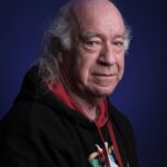 Een oudere man met lang grijs haar en een terugwijkende haarlijn draagt een zwarte hoodie met een kleurrijke afbeelding en een rode capuchon en poseert tegen een donkerblauwe achtergrond. Hij kijkt kalm naar de camera.