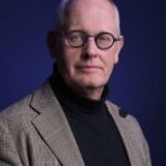 Een kale man met een ronde blauwe bril, een zwarte coltrui en een bruin geruite blazer poseert tegen een effen donkerblauwe achtergrond.