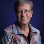 Een oudere vrouw met kort grijs haar en een bril kijkt naar de camera. Ze draagt een pastelkleurig jasje met patroon over een zwart topje, met een donkerblauwe achtergrond achter haar.