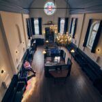 Een persoon zit alleen op een bank in een ruime kamer met hoge plafonds, donkerhouten meubels, kroonluchters, gewelfde ramen en een kleurrijk gebrandschilderd raam.