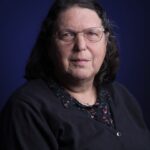 Een vrouw van middelbare leeftijd met bril en schouderlang donker haar, draagt een zwart vest over een blouse met patroon en poseert voor een portret tegen een donkerblauwe achtergrond.