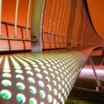 Een moderne voetgangersbrug bij nacht, gezien vanuit een lage hoek. De brug heeft een geperforeerd, verlicht oppervlak met gloeiende groene stippen en een gebogen metalen leuning. De lucht is oranje en beneden zijn lichtsporen van auto's zichtbaar.