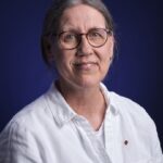 Een persoon met achterover gebonden grijs haar, een ronde bril en een wit overhemd met knoopsluiting, glimlacht zachtjes tegen een donkerblauwe achtergrond.