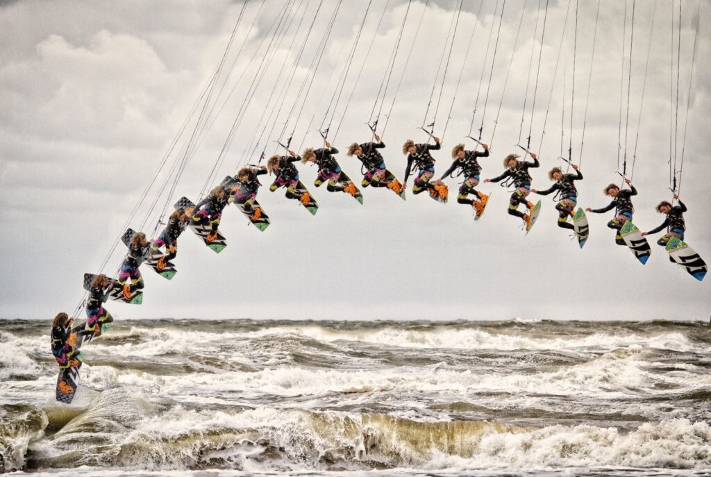 Een compositiefoto toont een persoon die kiteboardt, vastgelegd in meerdere beelden terwijl hij hoog boven de woeste oceaangolven springt en een boog vormt door de bewolkte lucht.