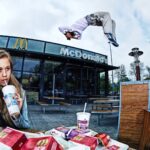 Een jonge vrouw zit buiten bij McDonald's, nippend aan een drankje en frietjes etend, terwijl een persoon een backflip uitvoert in de lucht boven het zitgedeelte buiten. Op de tafel staan verschillende etenswaren en drankjes.