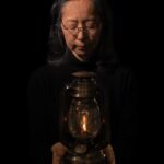 Een vrouw met een bril en lang donker haar houdt een brandende antieke lantaarn in beide handen en kijkt neer op de kleine vlam. Ze draagt een zwart topje tegen een donkere achtergrond.