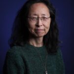 Een vrouw van middelbare leeftijd met lang, golvend zwart haar en een bril draagt een groene trui en een ketting met hanger en poseert tegen een donkerblauwe achtergrond. Ze heeft een kalme, neutrale uitdrukking.