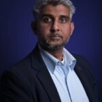 Een man met kort grijs haar en een baard, draagt een marineblauwe blazer over een lichtblauw overhemd, poseert tegen een donkerblauwe achtergrond. Hij kijkt recht in de camera met een neutrale uitdrukking.