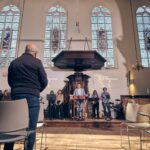 Een persoon staat op een podium in een kerk met gebrandschilderde ramen en spreekt een publiek toe. Muzikanten met instrumenten staan aan de zijkant en verschillende mensen staan op de voorgrond, met hun gezicht naar de spreker.
