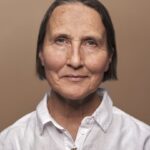 Een vrouw van middelbare leeftijd met kort bruin haar en blauwe ogen, die een wit overhemd met knoopjes draagt, kijkt kalm naar de camera tegen een effen beige achtergrond.