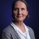 Een vrouw van middelbare leeftijd met kort bruin haar en een lichte huid, die een wit overhemd en een grijze trui draagt, poseert tegen een donkerblauwe achtergrond. Ze kijkt recht in de camera met een kalme, lichte glimlach.