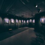 Een schemerige galerie met donkere gordijnen toont ingelijste portretfoto's van verschillende mensen op een rij langs de muren. Er staan een paar lege stoelen in de kamer.