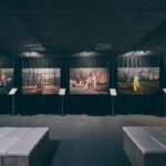Een zwak verlichte galerie toont vijf grote foto's op zwarte gordijnen, elk met afbeeldingen van mensen in verschillende binnen- en buitenomgevingen. Witte banken en informatiestands staan voor de kunstwerken.