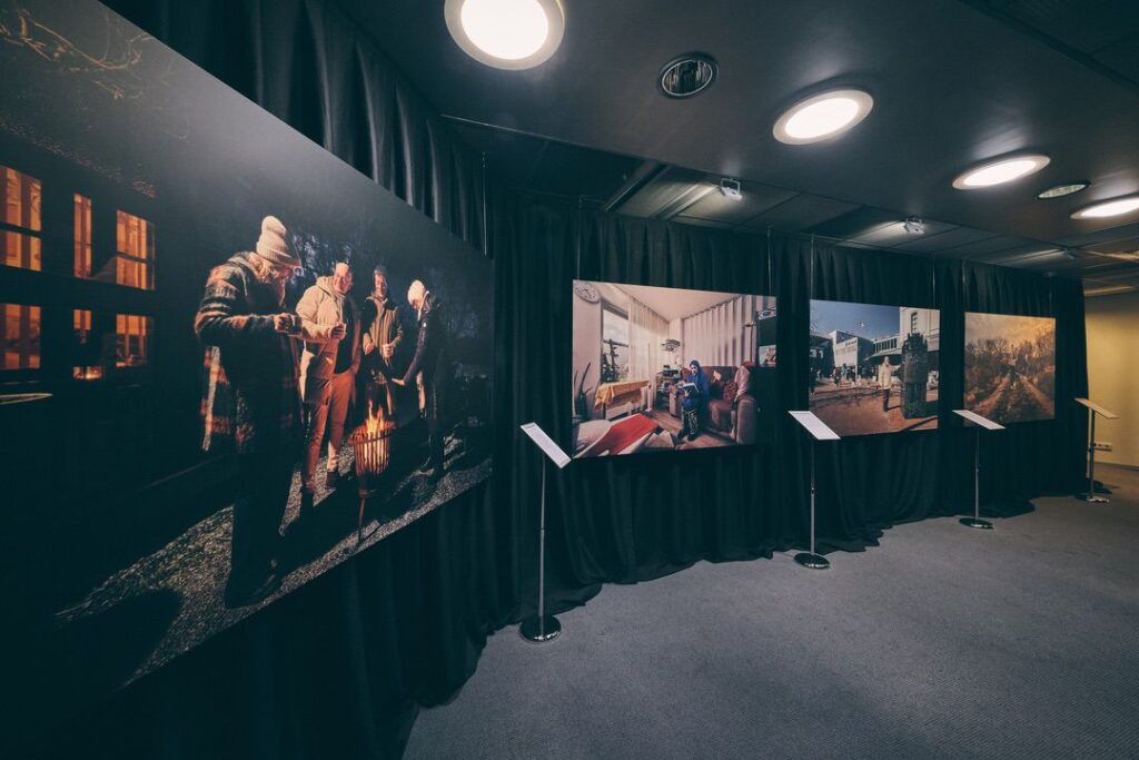 In een schemerige tentoonstellingsruimte hangen drie grote foto's op een zwart gordijn: mensen rond een vuur, een persoon in een gezellige woonkamer en een scène in de buitenlucht. Voor elke foto staat een informatiestandaard.