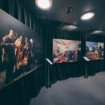 In een schemerige tentoonstellingsruimte hangen drie grote foto's op een zwart gordijn: mensen rond een vuur, een persoon in een gezellige woonkamer en een scène in de buitenlucht. Voor elke foto staat een informatiestandaard.