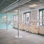 Een kunstinstallatie in een lichte kamer met honderden gevouwen papieren kraanvogels die in de lucht hangen tussen twee verticale palen. Grote ramen laten natuurlijk licht binnen en een spandoek met rode tekst staat tegen een blauwe muur.