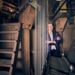 Een vrouw in een paarse blazer staat met gekruiste armen in een houten klokkentoren en kijkt naar grote metalen draden en machines naast een trap. Warme verlichting vult de historische, industriële ruimte.