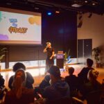 Een persoon spreekt op het podium voor een publiek, met een kleurrijk "Van de Straat" logo geprojecteerd op een scherm achter hen in een moderne indoor locatie met planten en spotlights.