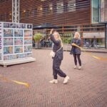 Twee vrouwen glimlachen en zwaaien met oranje linten terwijl ze buiten lopen in de buurt van een graffitikunstwerk, met fietsen en moderne gebouwen op de achtergrond.