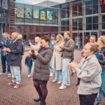 Een groep mensen, waaronder volwassenen en kinderen, staat buiten op een stenen plein te klappen en te kijken naar iets buiten beeld. Ze zijn gekleed in casual, warme kleding. Op de achtergrond zijn glazen gebouwen en kleurrijke kunstwerken te zien.