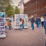 Mensen bekijken fototentoonstellingen in de open lucht op een stadsplein met grote fotopanelen. Sommige mensen staan te praten, terwijl anderen naar de displays kijken. Bomen en een modern gebouw staan op de achtergrond. Het lijkt een koele dag te zijn.