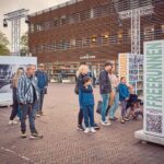 Mensen van verschillende leeftijden kijken naar borden in de buitenlucht met foto's en tekst. Op één bord staat "FREERUNNEN". De scène speelt zich af op een openbaar plein bij een modern houten gebouw.
