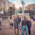 Een groep mensen maakt kennis met elkaar op een plein bij een theater. Sommigen staan te praten, terwijl een meisje op een fietsje rijdt. Grote posters en borden voor evenementen en shows zijn zichtbaar op nabijgelegen gebouwen.