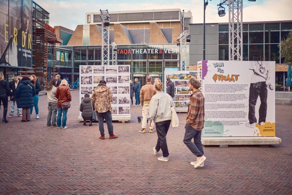 Mensen bekijken fotodisplays en informatieborden op een plein bij TAQA Theater De Vest. Het is een levendig tafereel met verschillende mensen die op een bewolkte dag door de exposities lopen en lezen.