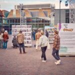 Mensen bekijken fotodisplays en informatieborden op een plein bij TAQA Theater De Vest. Het is een levendig tafereel met verschillende mensen die op een bewolkte dag door de exposities lopen en lezen.