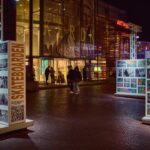 Nachtscène in de open lucht met mensen die in de buurt lopen van een helder verlicht gebouw met grote ramen. Op de voorgrond staan twee displays met skateboardfoto's en een QR-code, onderdeel van een tentoonstelling.