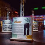 Nachtelijke openluchttentoonstelling met grote informatiepanelen, waarvan één met tekst en een zwart-witfoto. Moderne gebouwen met glazen gevels en kleurrijke lichten zijn zichtbaar op de achtergrond. Mensen lopen in de buurt.