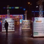 Een persoon staat 's nachts te kijken naar buitenpanelen met foto's en tekst op een felverlicht plein in de buurt van een theater, met kleurrijke lichten die de achtergrond verlichten.